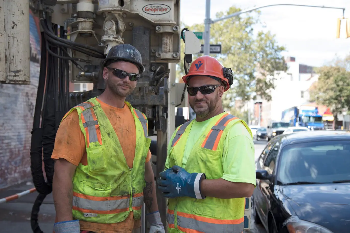 Two drillers wearing high visibility vests stand in front of a drill rig